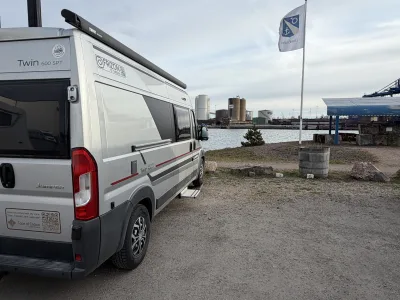 Frizze parkerad på ställplatsen vid Femöre Gästhamn med hamnen och flaggan i bakgrunden